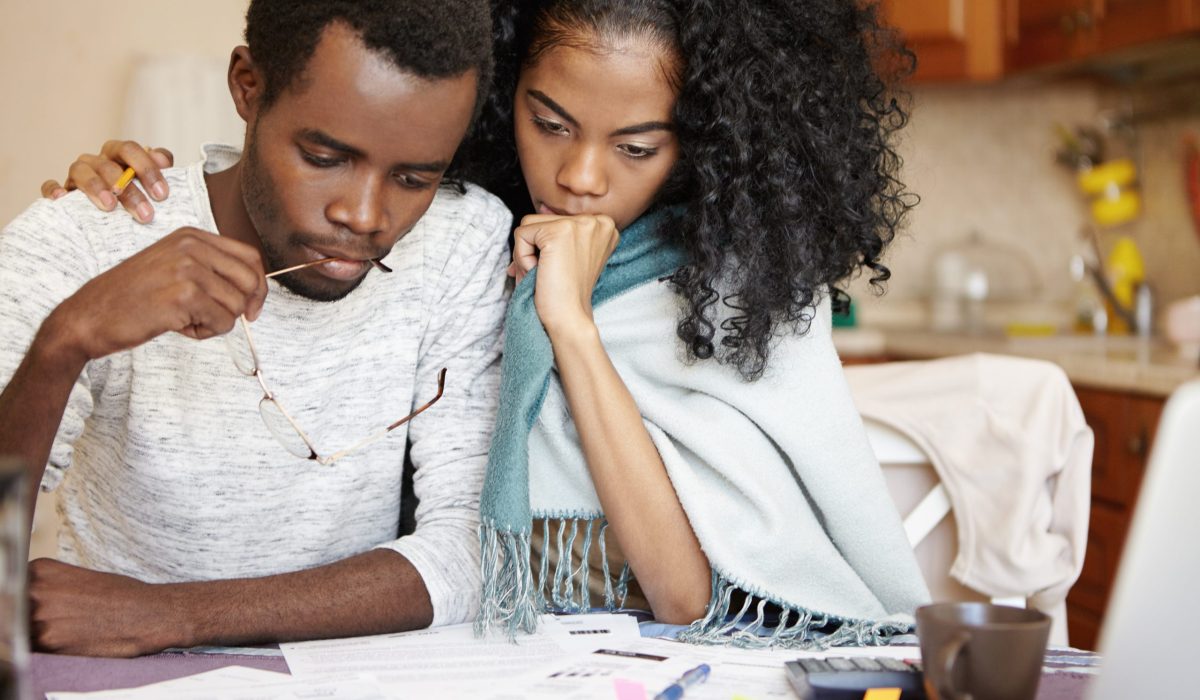 Frustrated young African couple having big problems paying utility bills: husband sitting over papers, holding glasses, having stressed and puzzled expression while his supportive wife embracing him